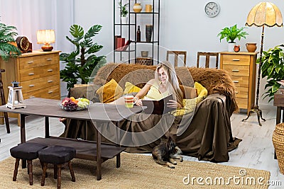 In the living room, a young woman is lying on the sofa reading a book. A dog is lying on the carpet. A multi-breed dog. Stock Photo