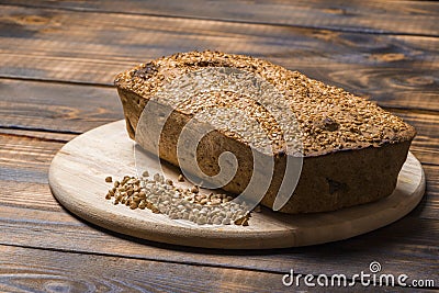 A loaf of freshly baked buckwheat bread lies on a round kitchen board on a brown wooden table. A handful of green Stock Photo