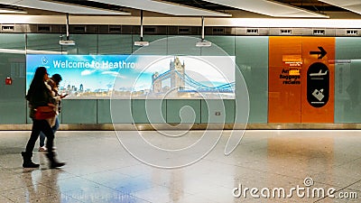 Two passengers walking past a a sign and image of Tower Bridge written Welcome to London at Heathrow Airport Terminal 3 Editorial Stock Photo