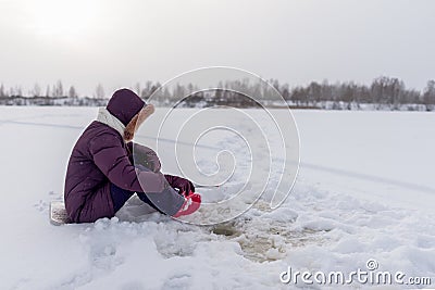 A lone angler girl is concentrating on catching fish with a winter fishing rod sitting on a substrate on the ice among flakes Stock Photo