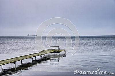 Lone bench on a thin boardwalk at sea and a faraway vessel amid winter gloominess covey sadness, solitude and desolation feelings Stock Photo