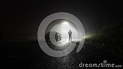 A lone car, parked on the side of the road, underneath a street light, with a hooded figure, on a spooky, scary, rural, country Stock Photo