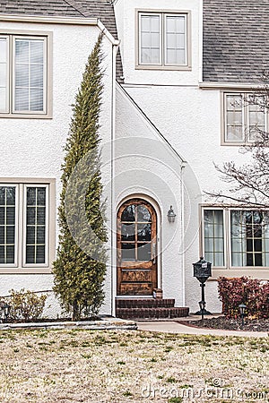 Lone cypress tree grows by arched wooden front door of charming white stucco house with angles and standing iron mailbox Stock Photo