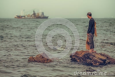 Lone fisherman gazes out at large fishing boat. Editorial Stock Photo