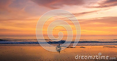 A lone motorcycle and rider driving along a beach at sunset Stock Photo