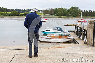 A lone retired male standing on the edge of a quay and looking out over the river and the boats that are moored up close by Editorial Stock Photo