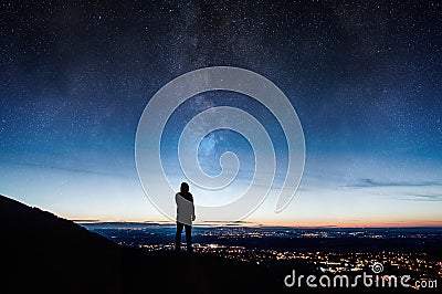 A lone silhouetted hooded figure. Standing on a hill looking down on city lights at night with a galaxy and stars rising in the ni Stock Photo