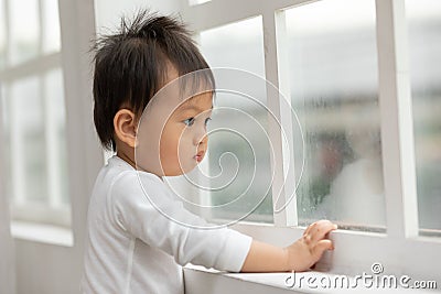 Lonely Adorable asian baby toddler standing in front of windows and looking outside waiting mom or dad come back home.Sad Baby Stock Photo