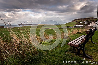 Lonely bench in a cold and windy coastal city, pointing to the sea with dramatic cloudy sky while grass is blown by the wind. Stock Photo