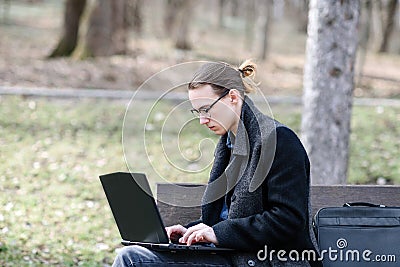 A lonely European student with glasses and a coat works on a laptop while sitting on a park bench Stock Photo