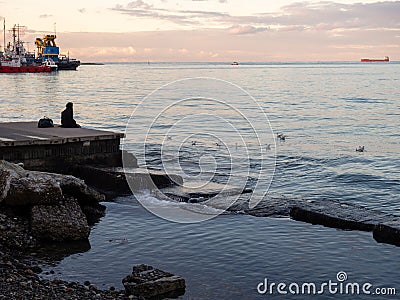 A lonely girl sits on a pier in the port. Sitting on the edge of the water Stock Photo