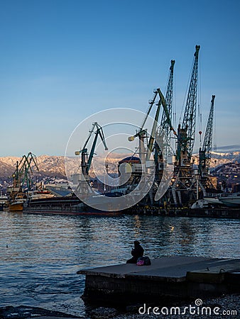 A lonely girl sits on a pier in the port. Sitting on the edge of the water. Uses a phone Stock Photo