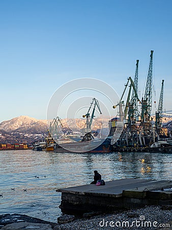 A lonely girl sits on a pier in the port. Sitting on the edge of the water. Uses a phone Stock Photo