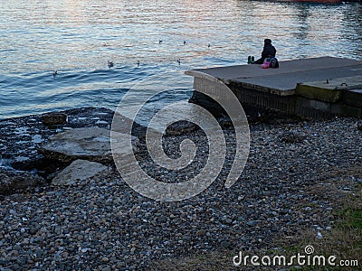 A lonely girl sits on a pier in the port. Sitting on the edge of the water. Uses a phone Stock Photo