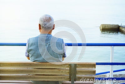 Lonely gray-haired elderly man sitting by the sea on a bench, a view from the back, contemplation of nature and a relaxing holiday Stock Photo