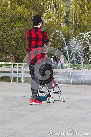 Lonely single father hipster in checkered red and black shirt with a stroller walking in the city park Editorial Stock Photo