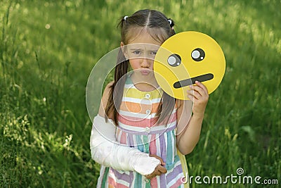 Lonely upset girl with broken arm and a cast holds crying emoticon Stock Photo
