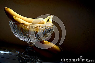 Ripe bananas in a crystal fruit vase salad bowl in sunbeam in dark shadows Stock Photo