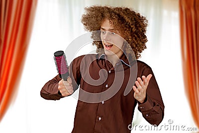 A long-haired curly-haired guy in a brown shirt uses a metal round comb. Emotions before a haircut in a hairdresser Stock Photo