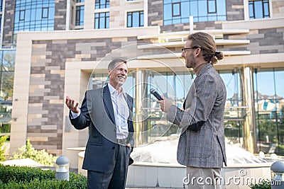 Long-hareid male journalist in eyeglasses interviewing smiling male in suit outside Stock Photo