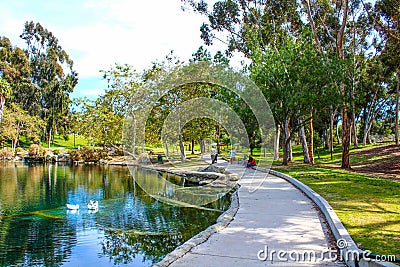 A long paved footpath along the lake surrounded by lush green trees, plants and grass with people walking along the footpath Editorial Stock Photo