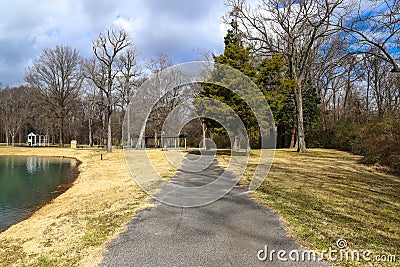 A long rocky footpath with a brown wooden pergola at the end with yellow winter grass in the park surrounded by lush green trees Stock Photo