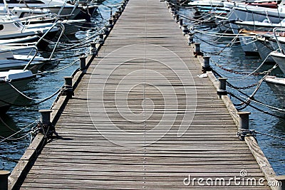 Long wooden pier with dense iron mooring bollards on each side and multiple small boats tied with strong ropes and chains Stock Photo