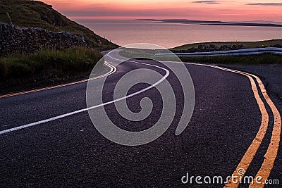 Looking down a sweeping road near the Cliffs of Moher to beautiful pink sky sunset over the island of Inisheer Stock Photo