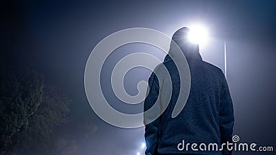 Looking up from behind at a mysterious, moody, hooded figure. On a foggy winters night, with street lights behind Stock Photo