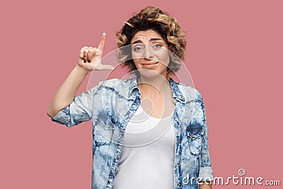 Loser. Portrait of sad young woman with curly hairstyle in casual blue shirt standing with loser gesture and looking at camera Stock Photo