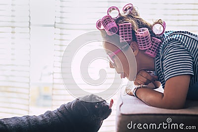 Love in funny moment with woman lay down on the sofa with pink curlers on hair and black lovely pug dog kissing her or making nice Stock Photo