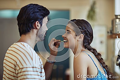 Love makes everything taste better. Shot of an affectionate young couple snacking while preparing a meal in their Stock Photo