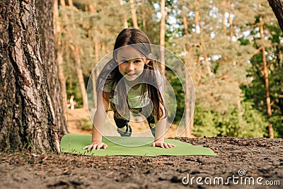 Loveable girl with flowing hair doing plank exercise on green mat outdoor. Junior female athlete working out in nature. Stock Photo