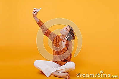 Lovely barefooted girl taking picture with peace sign. Romantic lady in white pants sitting with legs crossed and making Stock Photo