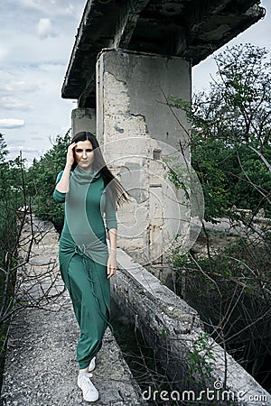 Lovely girl in green dress and with loose hair goes through the old stone bridge in the out of town. Stock Photo