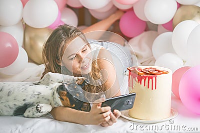 Lovely lady in pajama making selfie in her bedroom using phone and play with her dog. Indoor portrait girl with baloons in Stock Photo