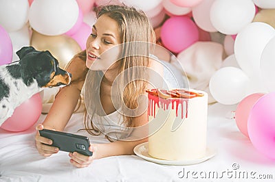 Lovely lady in pajama making selfie in her bedroom using phone and play with her dog. Indoor portrait girl with baloons in Stock Photo