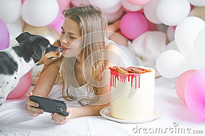 Lovely lady in pajama making selfie in her bedroom using phone and play with her dog. Indoor portrait girl with baloons in Stock Photo