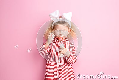 Lovely little girl in a striped red and white dress and bunny ears on her head inflates soap bubbles standing against a Stock Photo