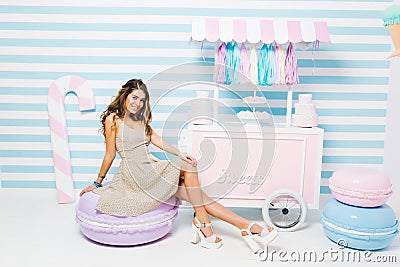 Lovely tanned girl wearing vintage dress sitting on purple chair looking like macaroon near the sweets counter. Indoor Stock Photo