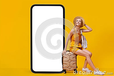 Lovely young blonde woman sitting on suitcase near giant smartphone with mockup, booking summer vacation online Stock Photo