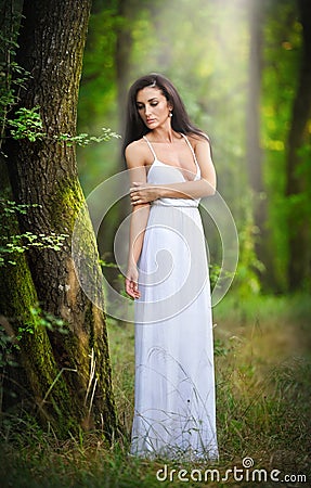 Lovely young lady wearing an elegant long white dress enjoying the beams of celestial light on her face in enchanted woods. Long Stock Photo