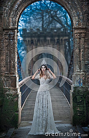 Lovely young lady wearing elegant white dress and silver tiara posing on ancient bridge, ice princess concept. Pretty brunette Stock Photo