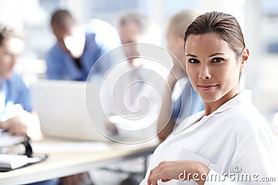 She loves her job. A beautiful medical professional looks at the camera with colleagues working in the background. Stock Photo