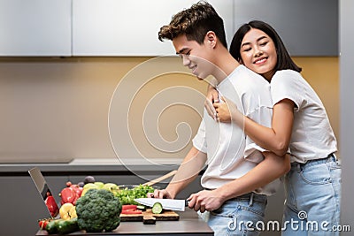 Asian Wife Hugging Husband While He Cooking Dinner In Kitchen Stock Photo