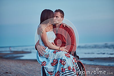 Loving couple, a woman sits on her husband`s lap, resting on a beach against a background of a bright dawn. Stock Photo