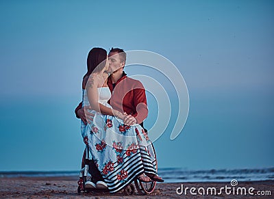 Loving couple, a woman sits on her husband`s lap, resting on a beach against a background of a bright dawn. Stock Photo