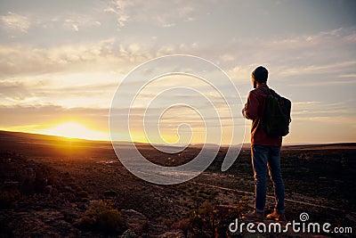 Low angle view of young male hiker with backpack standing on mountain top while hiking looking at sunrise Stock Photo