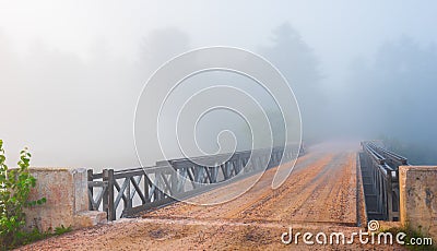 Low cloud in bright mist. single lane road, steel & timber bridge into vanishing forest. Stock Photo