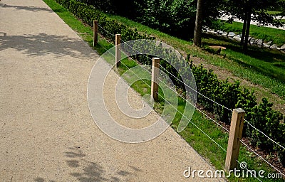 A low fence of land with three steel ropes and wooden poles separates the gravel path from the lawn and the flowerbed with boxwood Stock Photo